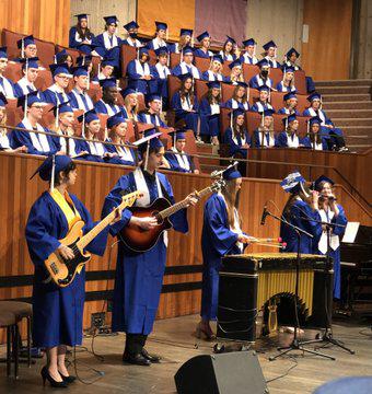 A group of graduates in caps and gowns perform a song, all playing various instruments. 